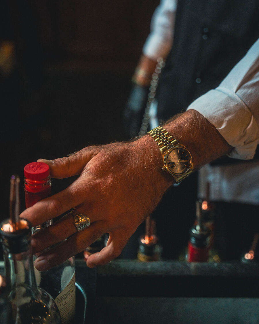Gold watch and ring reaching for a bottle behind the bar